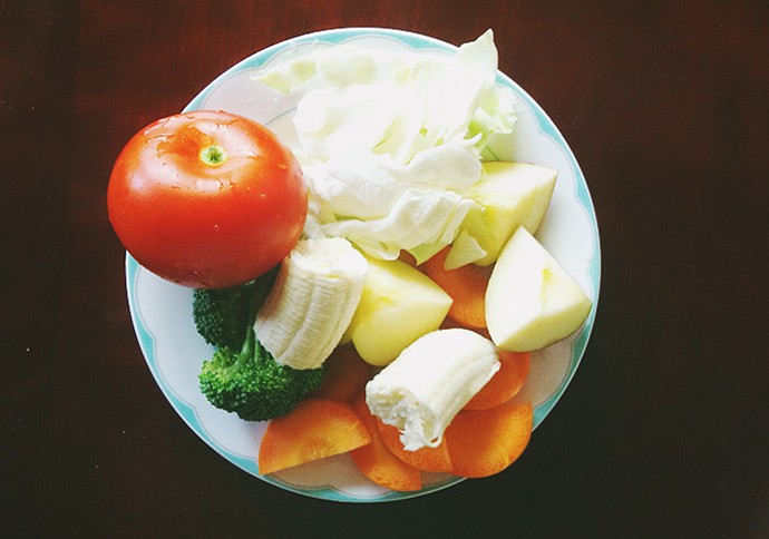 접시 위에 양배추, 사과, 당근, 바나나, 브로콜리가 잘라져 있다. 토마토는 잘리지 않은 온전한 모양이다