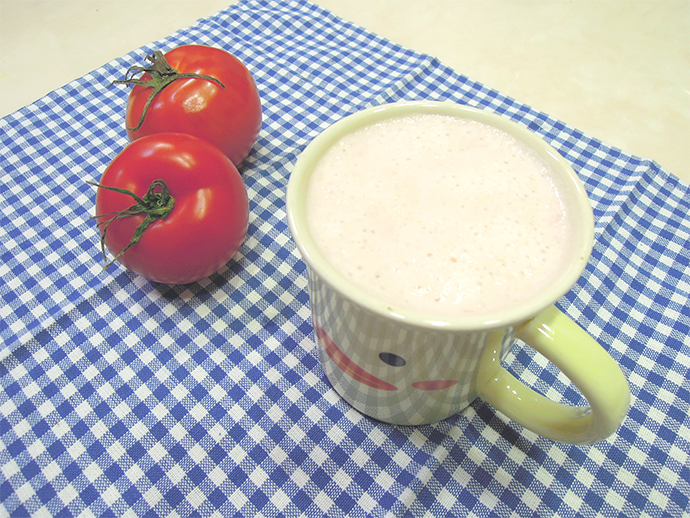 토마토 우유가 노란색 병아리가 그려진 컵에 담겨있다. 컵 왼쪽으로는 빨간 토마토 2알이 놓여있다.