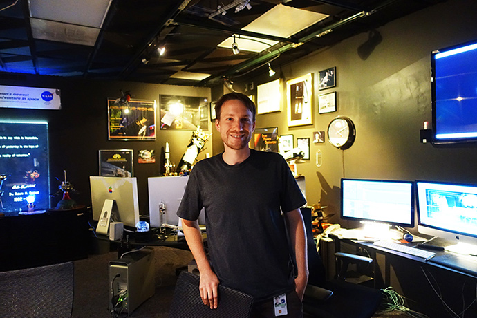 어두침침한 스튜디오에 영상디자이너 Brian이 서 있다. 그 뒤로는 우주 관련 피규어들이 보인다.