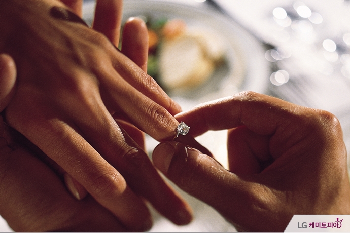 한 남성의 손이 다른 여성의 손가락에 다이아몬드 반지를 끼워주고 있다.