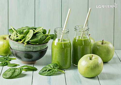 녹색 사과, 채소가 있고, 그 옆에 투명한 유리병에 녹색주스가 담겨있다. 